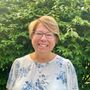 Woman with short blonde hair and glasses smiles in front of leafy green bushes; she wears a white floral blouse.