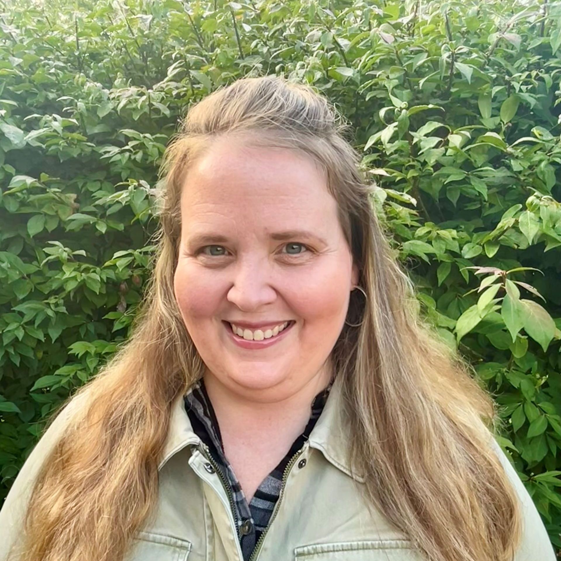 Woman with long blonde hair, smiling at the camera, wearing a green jacket, in front of leafy green plants.