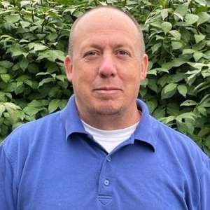 Man in blue polo shirt, white undershirt, looking at the camera. Green foliage background.