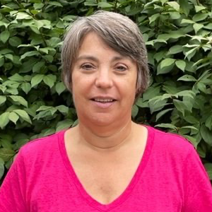 Woman with short gray hair and a pink shirt smiles in front of green foliage.