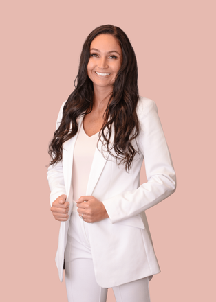 A woman in a white suit is standing in front of a pink background.