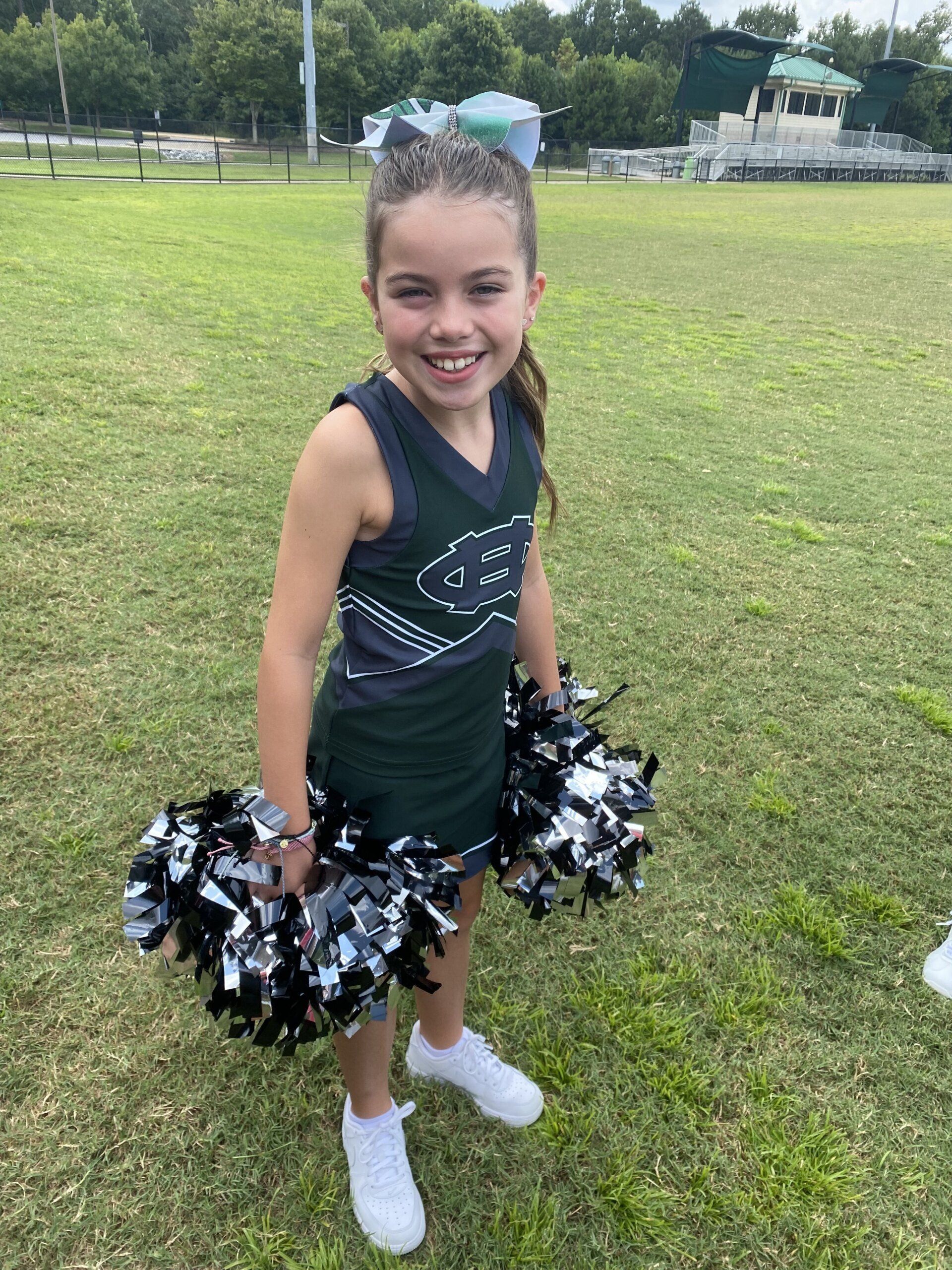 A young girl in a cheerleading uniform is holding pom poms in a field.