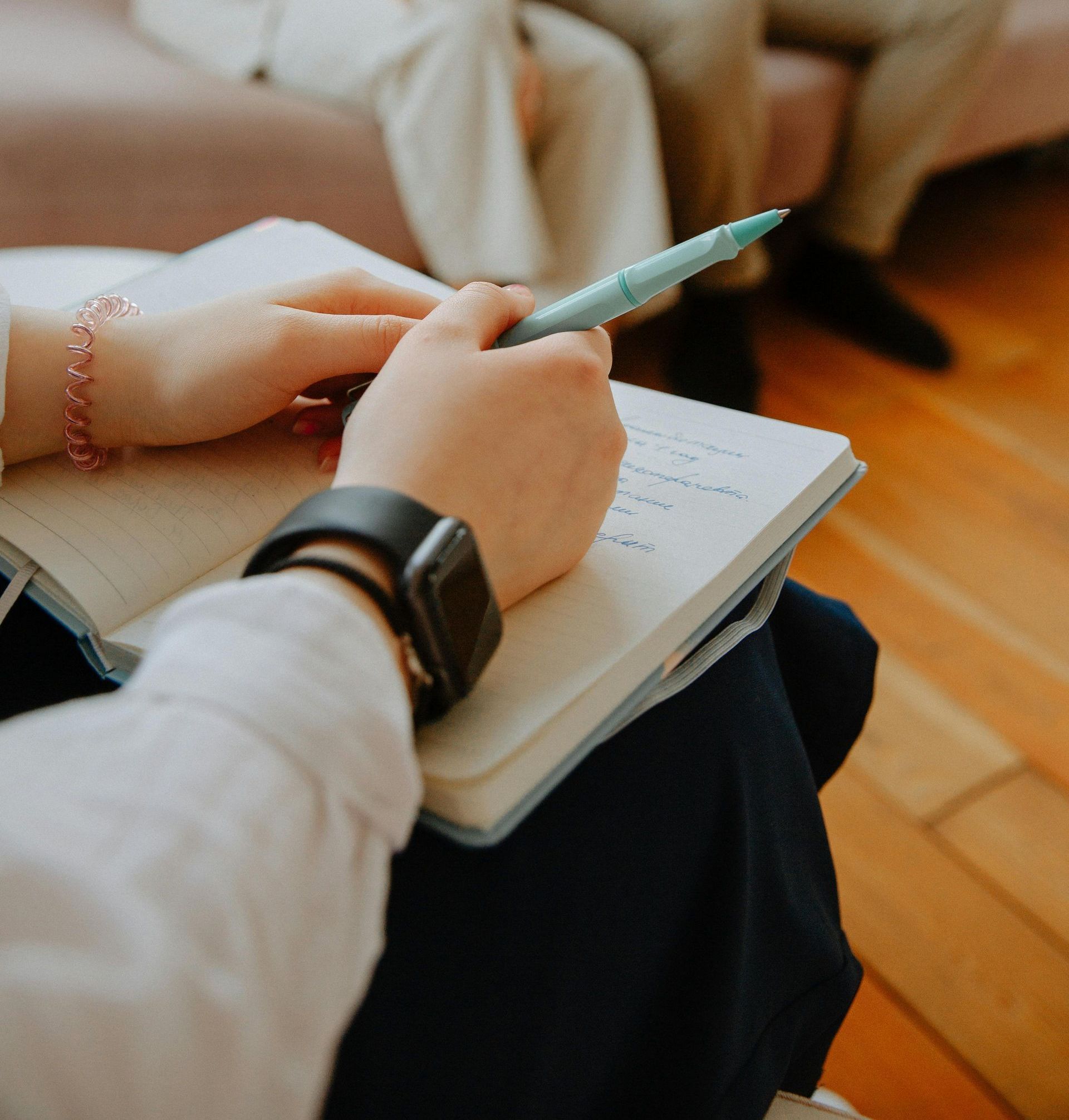 Hands holding a pen and notebook on a couch, with a smartwatch visible on the wrist.