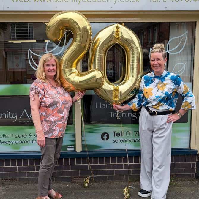 Two people standing beside large gold “20” balloons in front of a shop window.