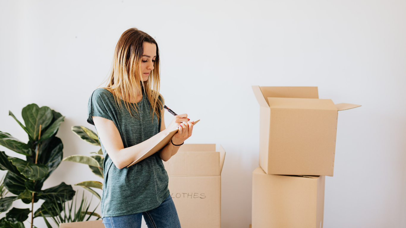 Person writing on a clipboard beside stacked moving boxes in a bright room