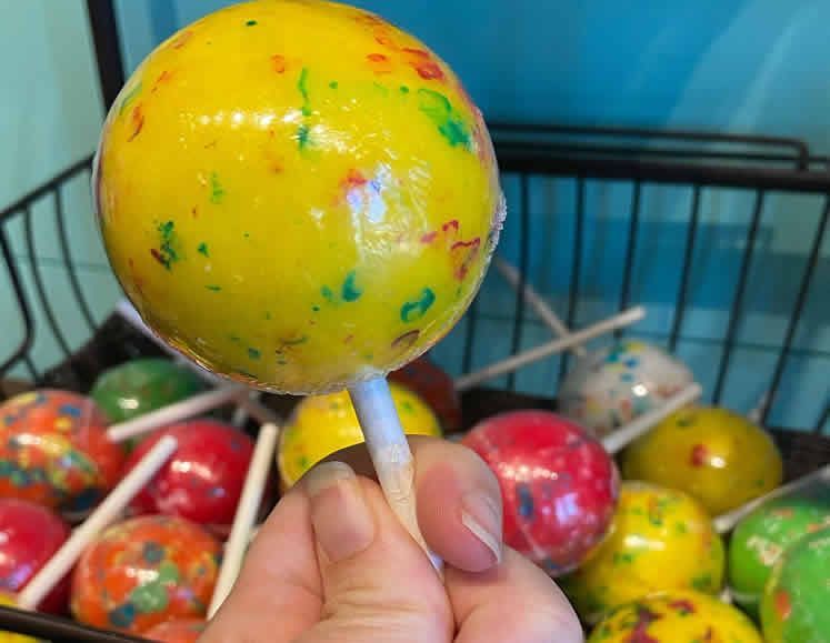 A person is holding a yellow lollipop in front of a basket of lollipops.