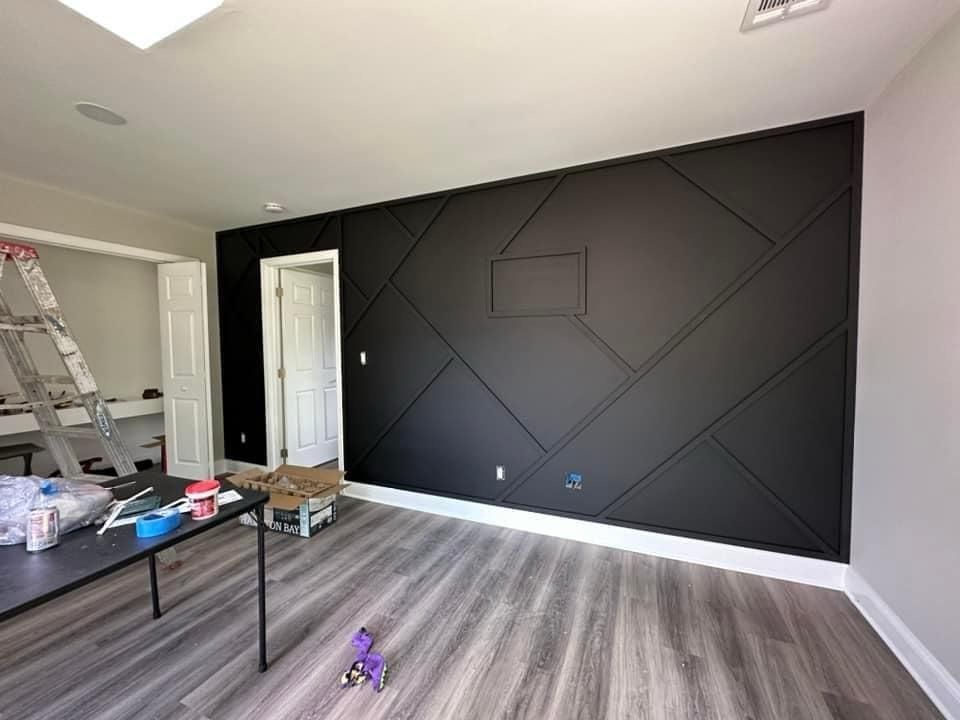 A black geometric accent wall with a rectangular opening, next to a doorway and white trim. Gray flooring and a ladder present.