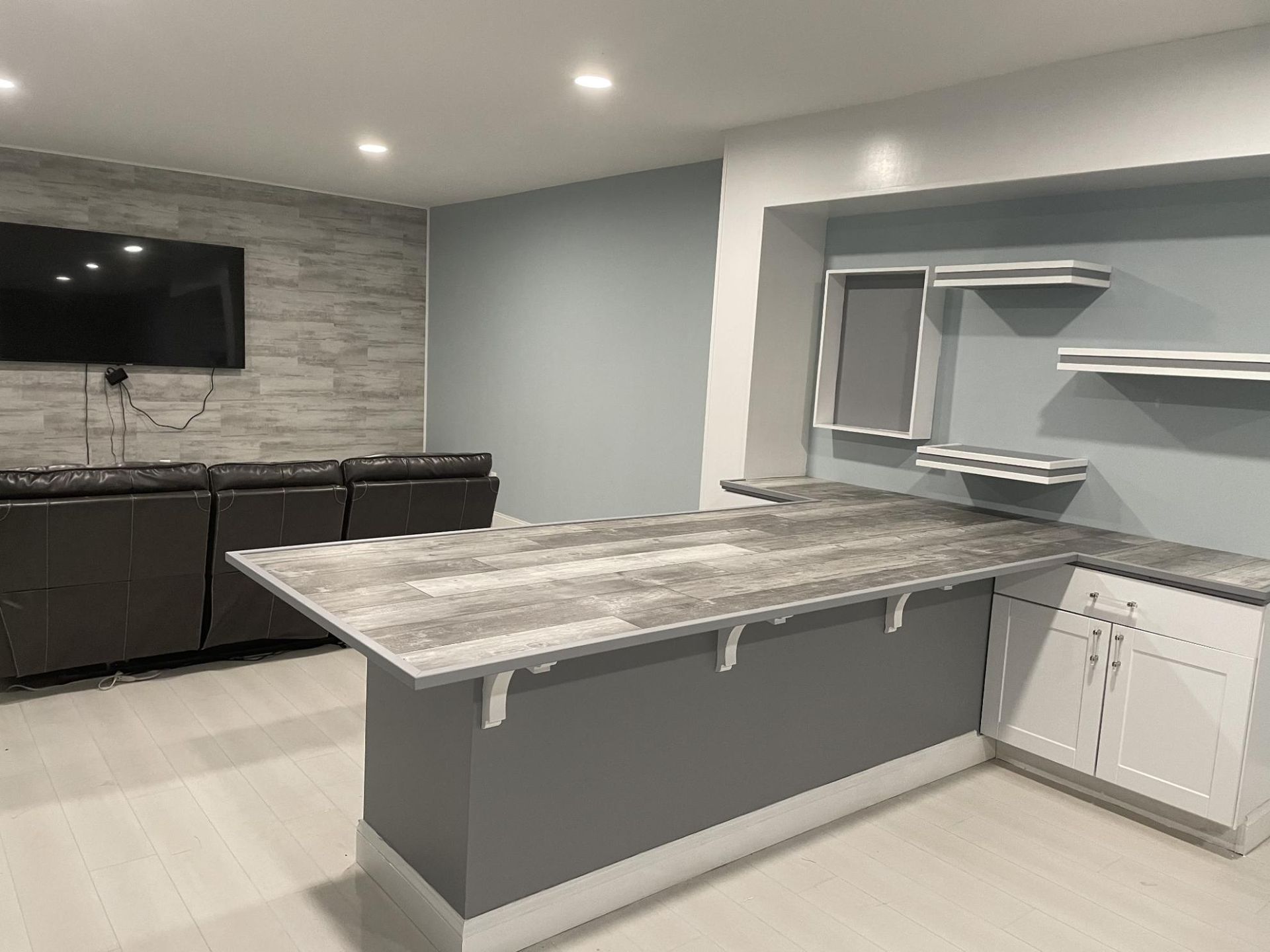 Basement bar area with gray countertop, white cabinets, and built-in shelves on a light blue wall.