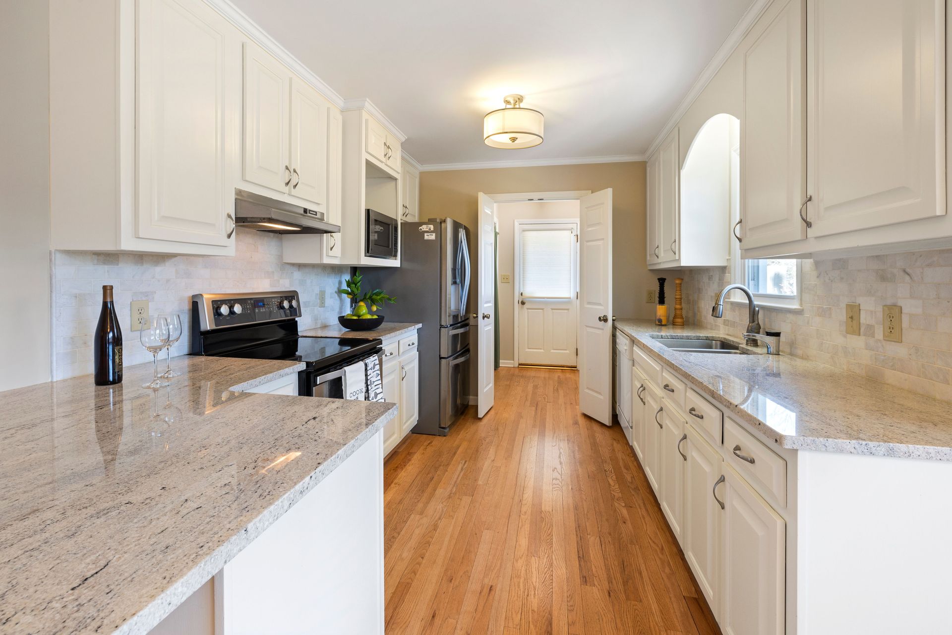 Bright galley kitchen with white cabinets, granite counters, stainless appliances, and wood floors