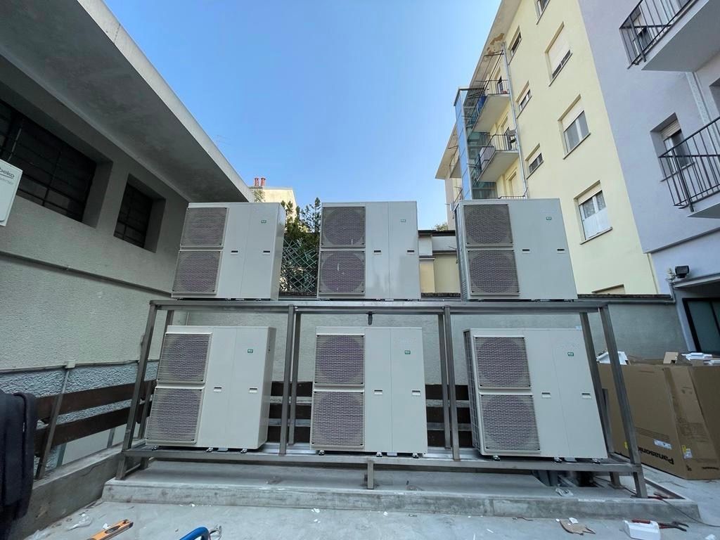 Sei unità esterne di climatizzazione di colore grigio chiaro, montate su una struttura metallica tra due edifici, sotto un cielo azzurro e limpido.
