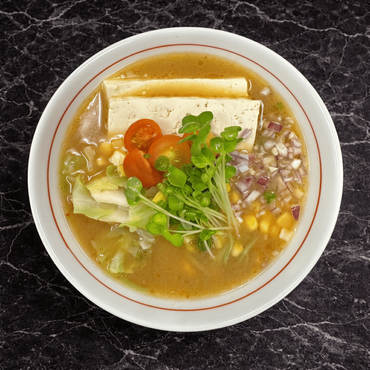 A bowl of ramen with tofu, corn, mushrooms, cabbage, and green onions in a broth, set on a dark, marbled surface.