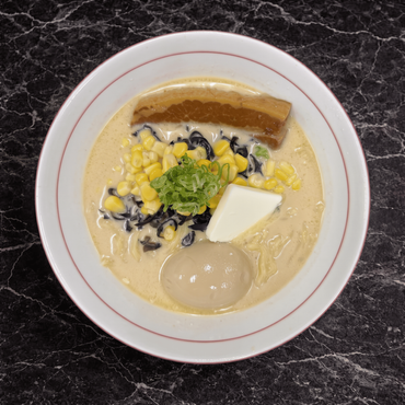 A bowl of ramen with pork belly, a soft-boiled egg, corn, bamboo shoots, butter, and green onions in a creamy broth.
