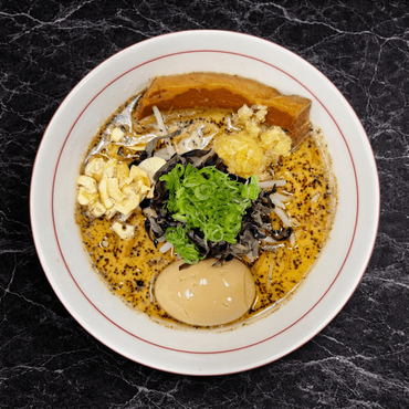 A bowl of ramen with pork slices, a soft-boiled egg, corn, green onions, and fried garlic in a dark, savory broth.
