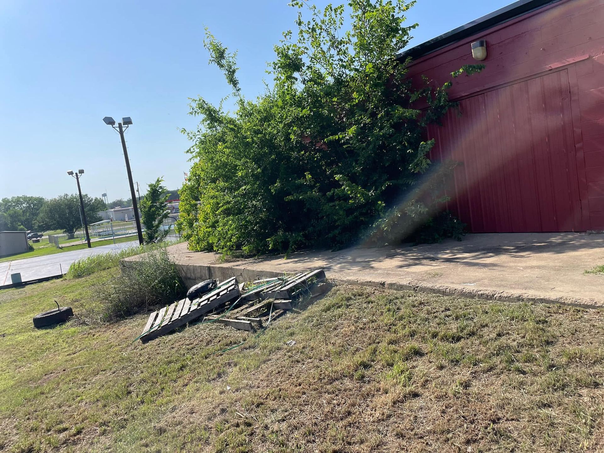 Broken chain-link fence beside a red building, with debris on a grassy slope near a parking lot.