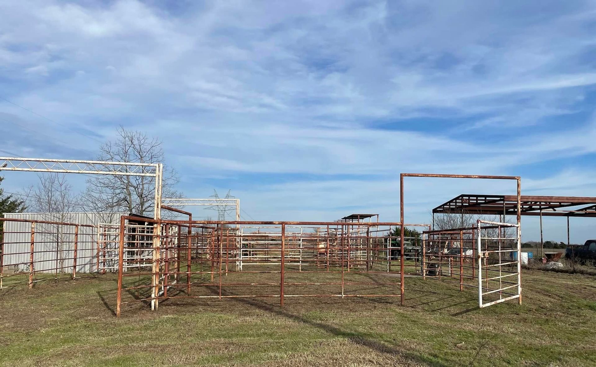 Rusty metal livestock pens in a grassy field under a blue sky, with a small shelter and white shed nearby.