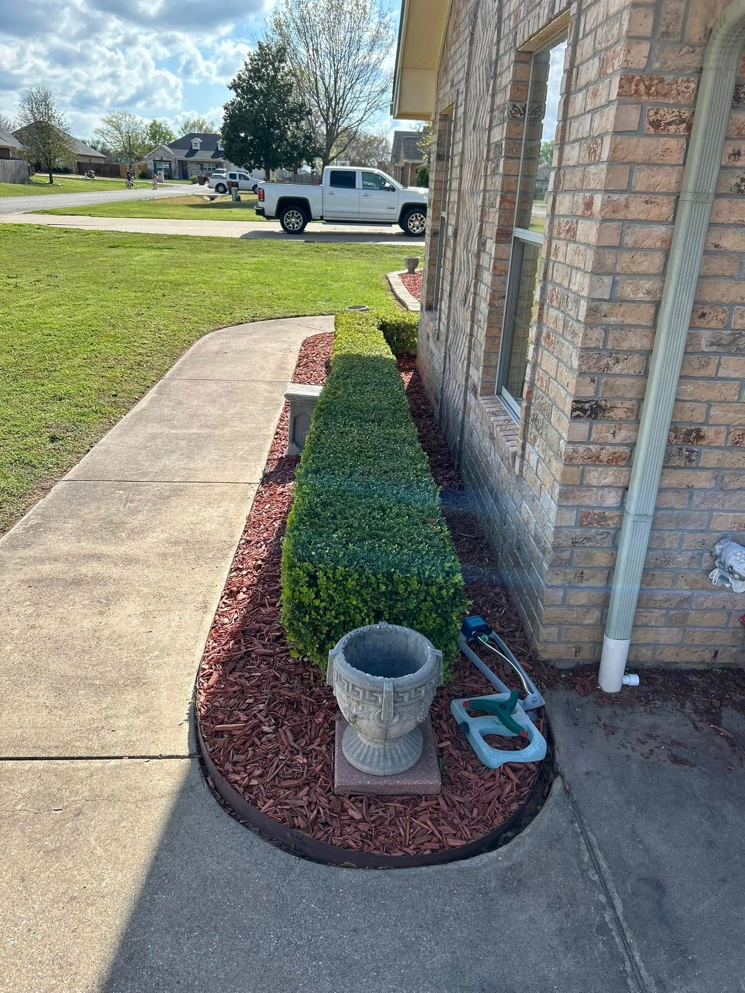 Front yard flower bed beside a brick house, with red mulch, green shrubs, and a stone planter by the walkway.