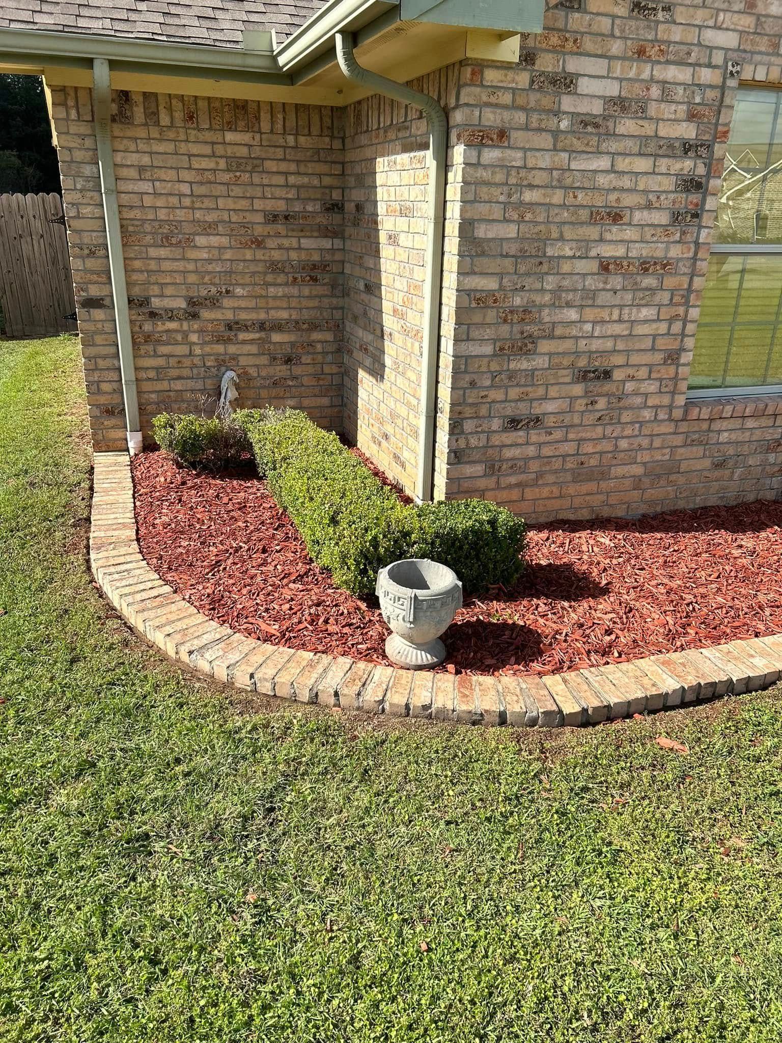 Brick house corner with red mulch bed, trimmed shrubs, and a small stone garden light along the lawn edge