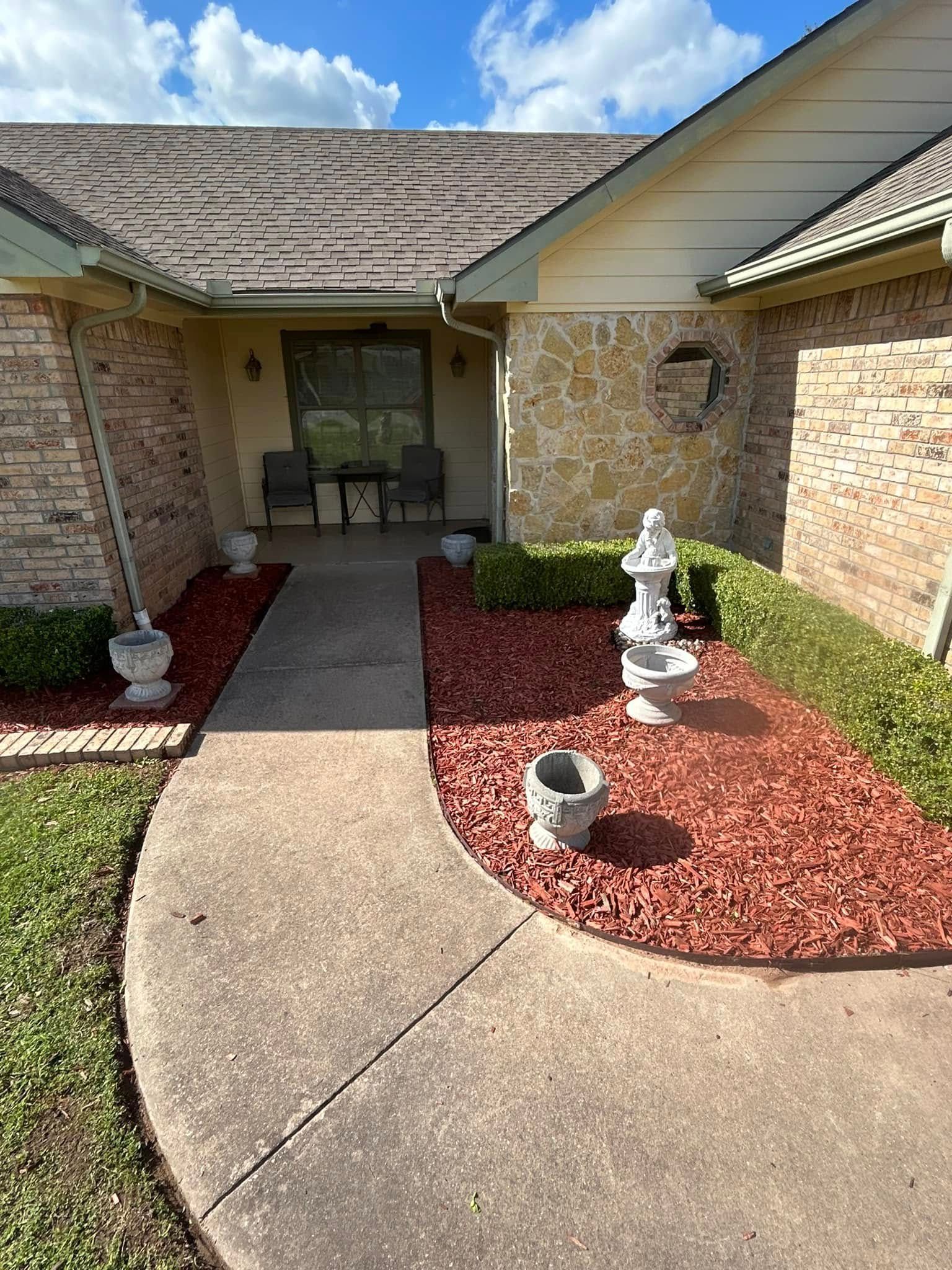 Front entrance walkway with brick house, red mulch beds, and small white garden statues under a covered porch