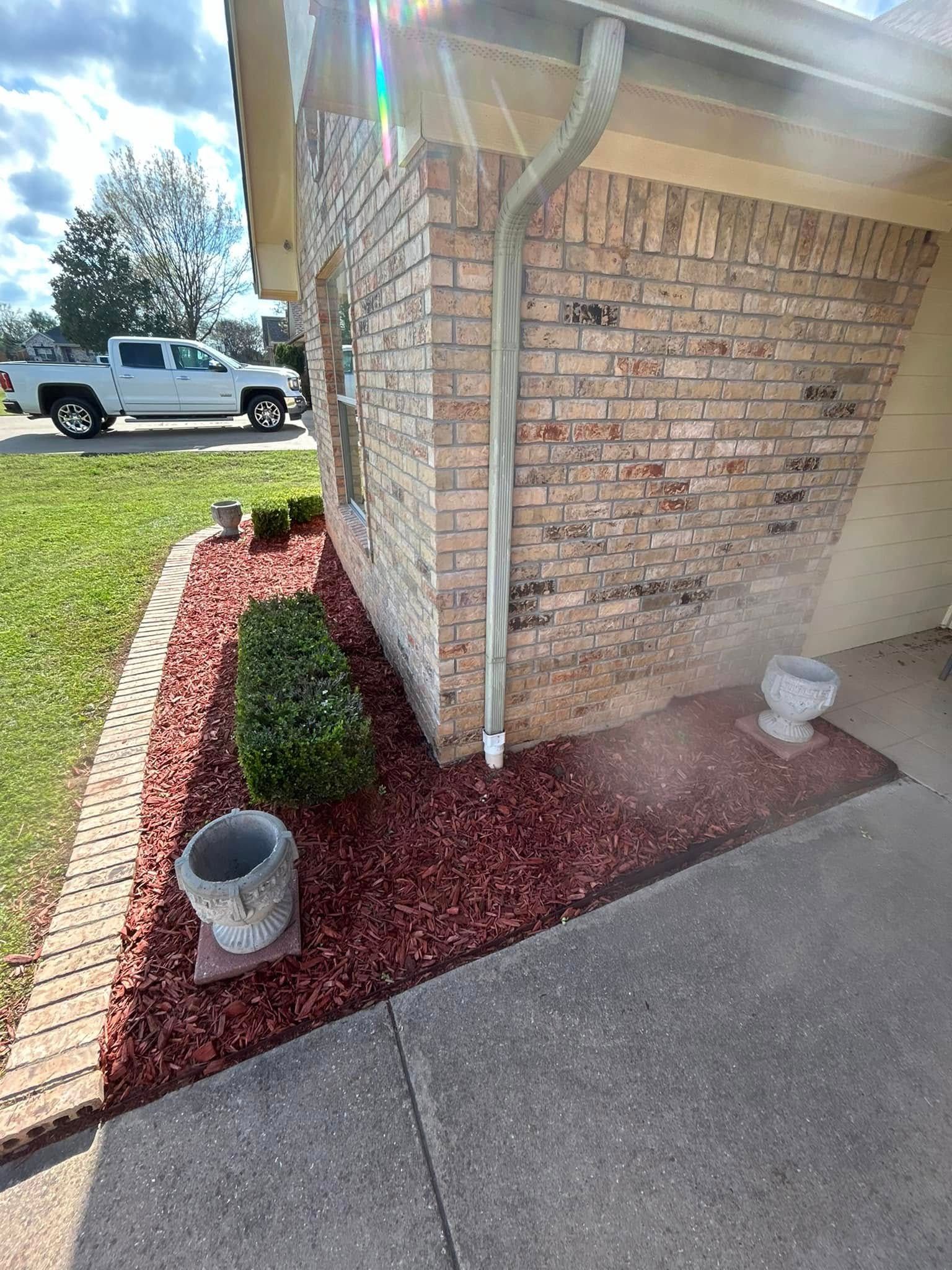 Brick house corner with red mulch bed, small shrubs, and a white downspout beside a driveway