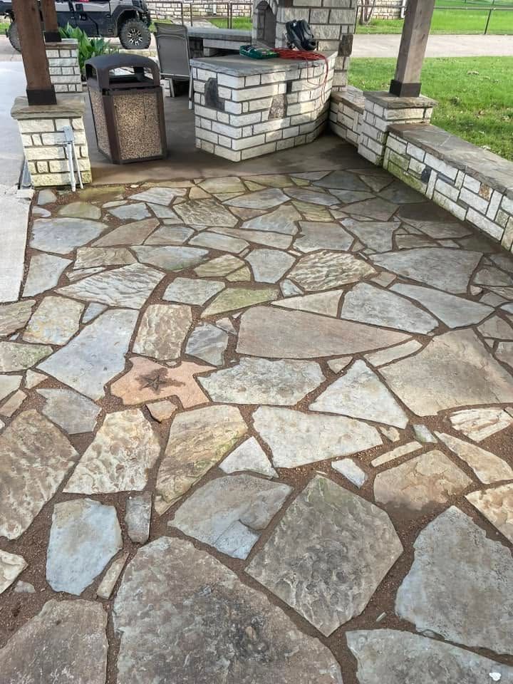 Stone-paved park patio with brick planters and a trash can beside a grassy area