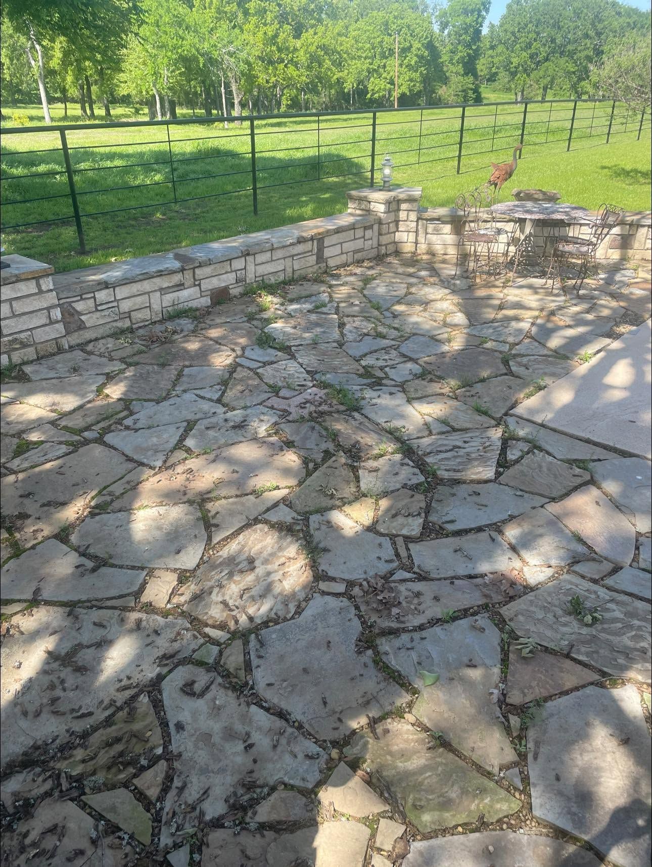 Stone patio with cracked paving beside a low stone wall and green grassy yard in sunlight