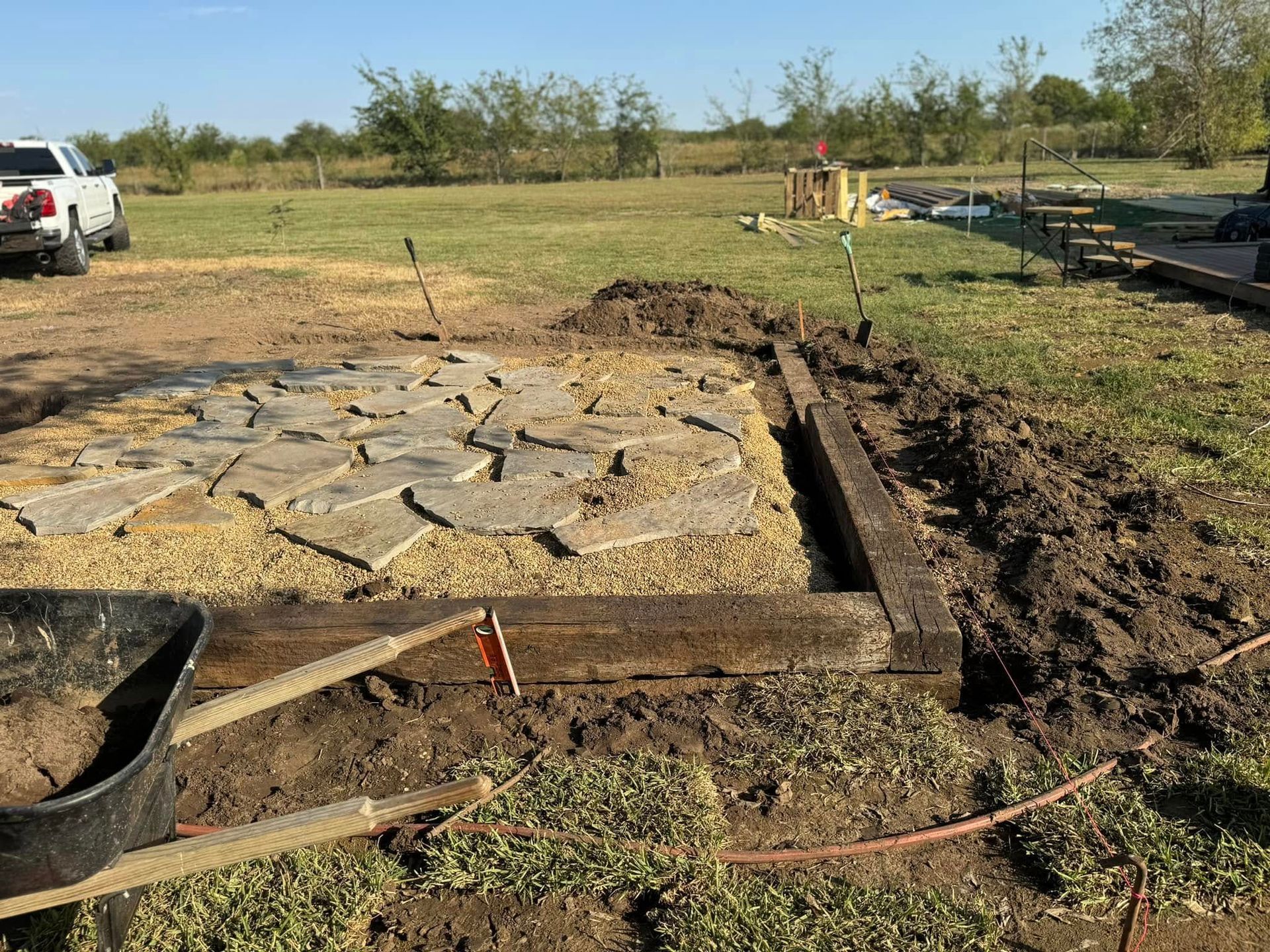 Stone foundation under construction in a grassy field, with dirt trenches and tools around it