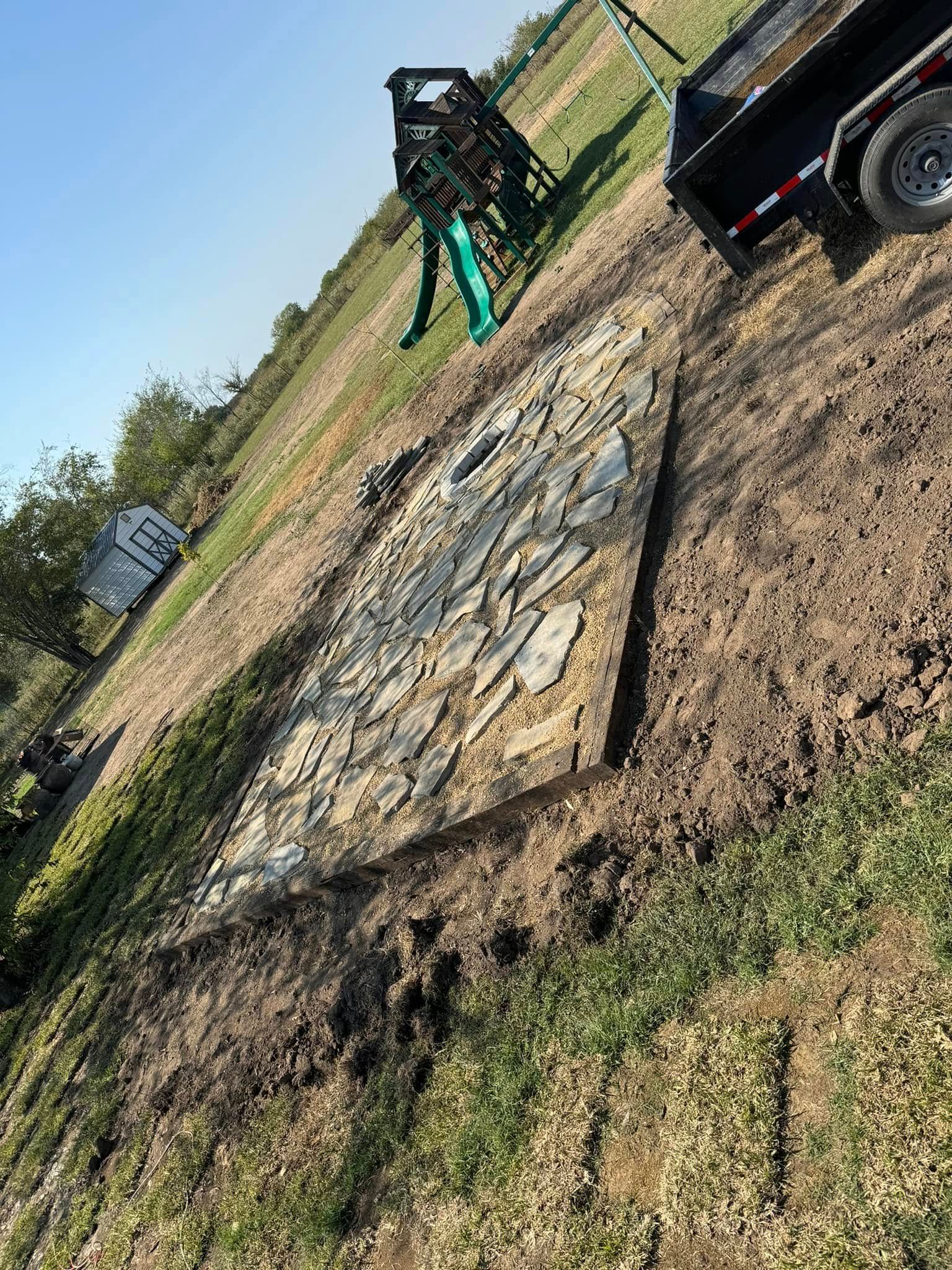 A slanted stone patio under construction beside a parked ATV in a grassy field.