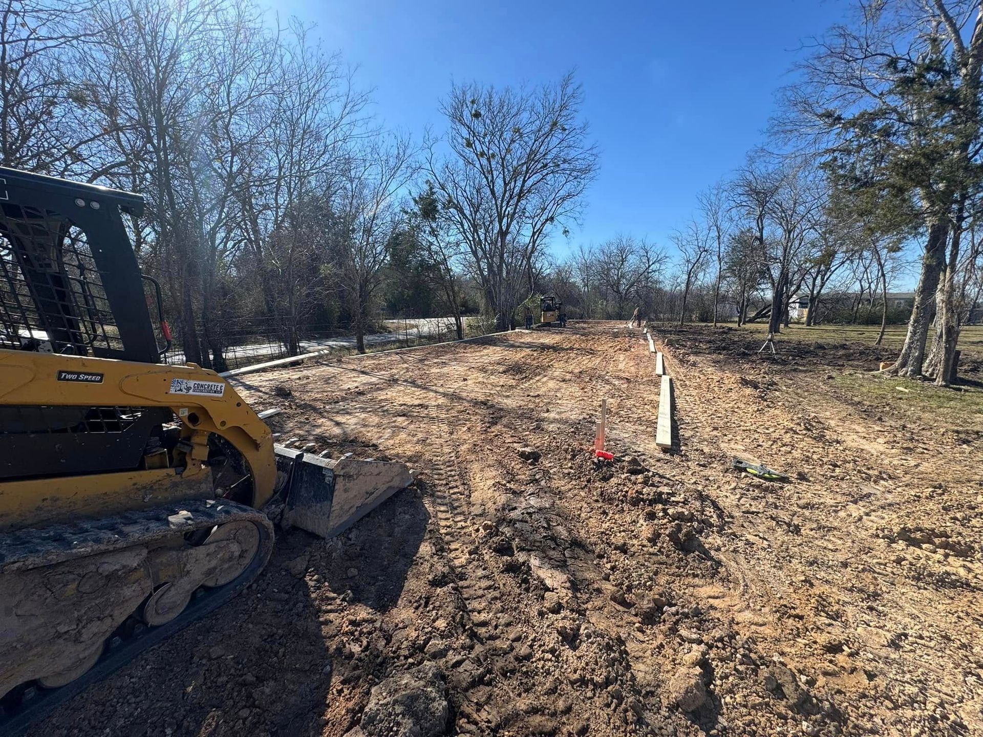 Excavator working on a dirt clearing with trees and a white post under a blue sky