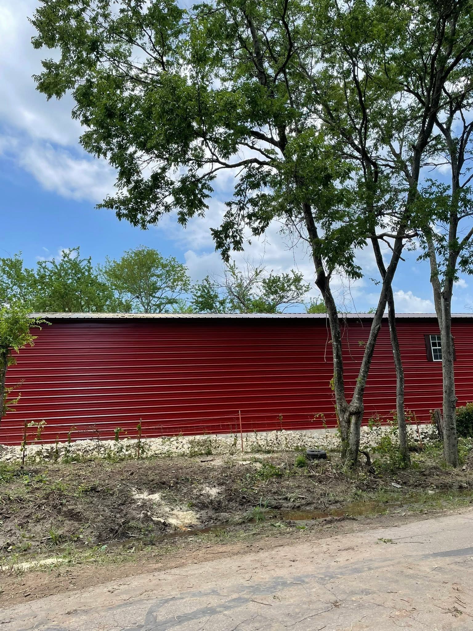 Red corrugated building behind trees under a blue sky, beside a dirt road.