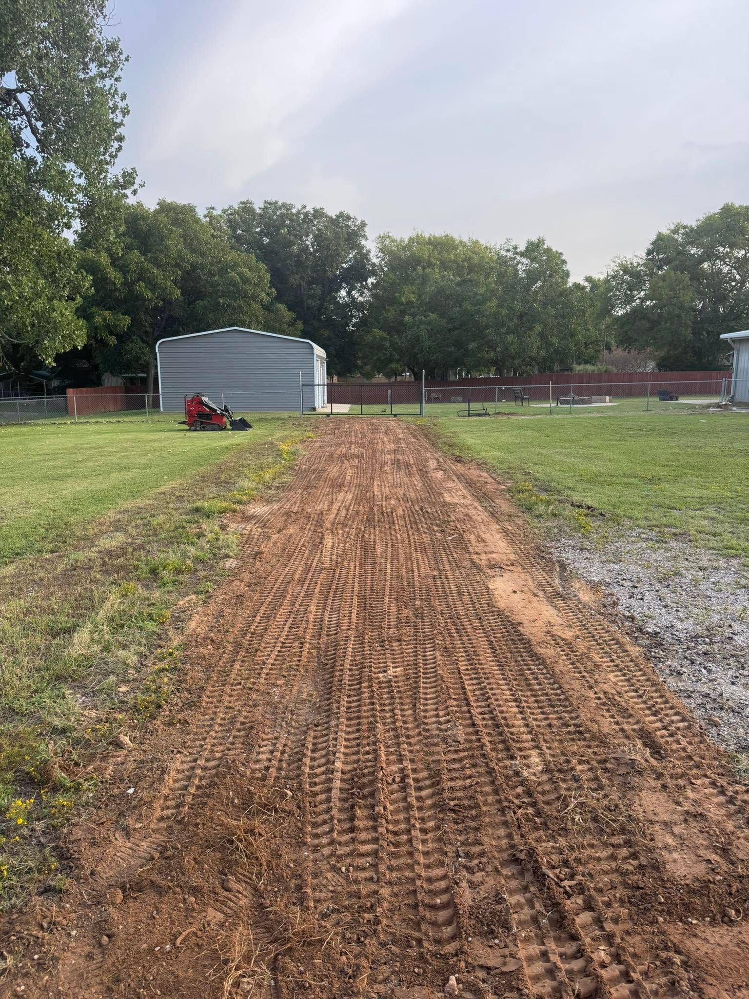 Gravel path through a grassy yard toward a gray metal shed, with trees in the background and a red tractor nearby