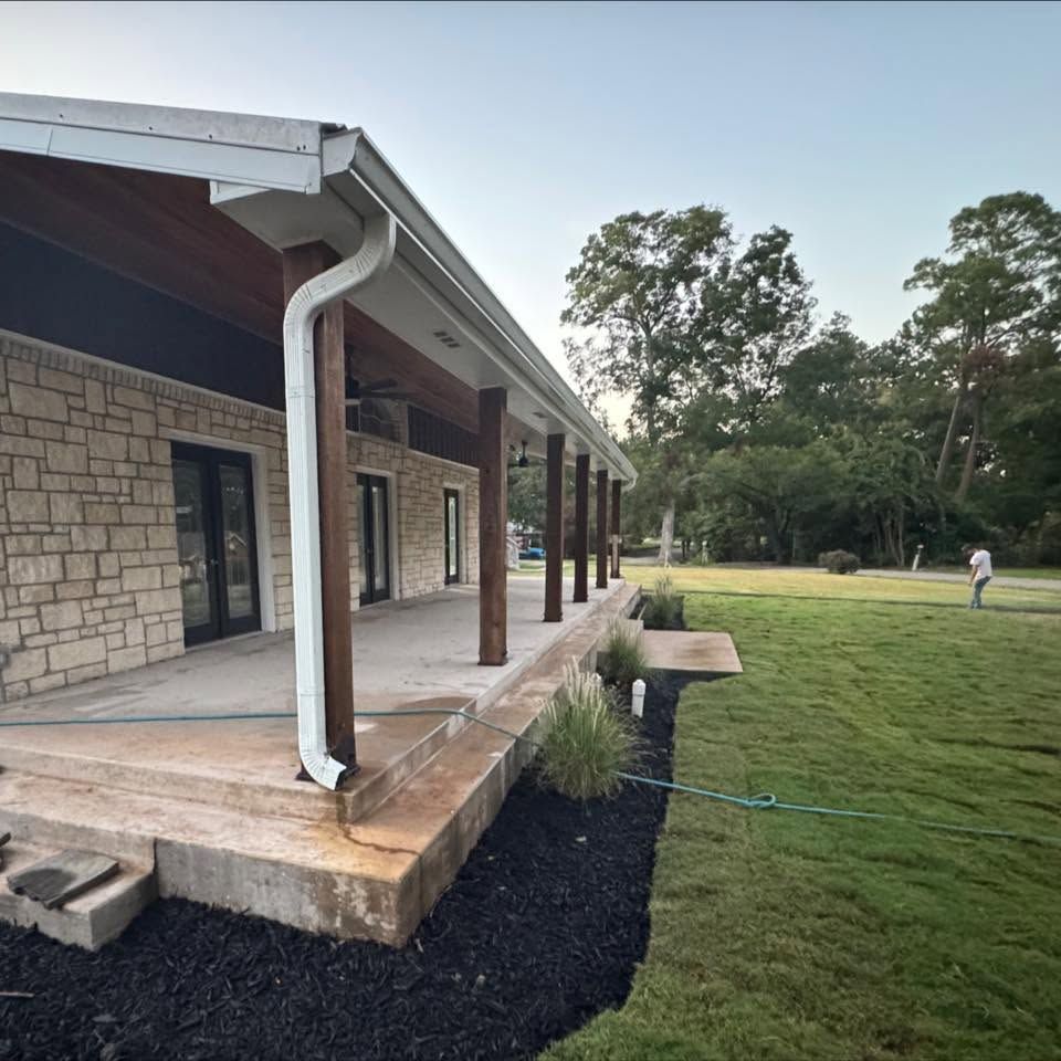 Covered stone patio beside a grassy yard, with white gutters and trees in the background