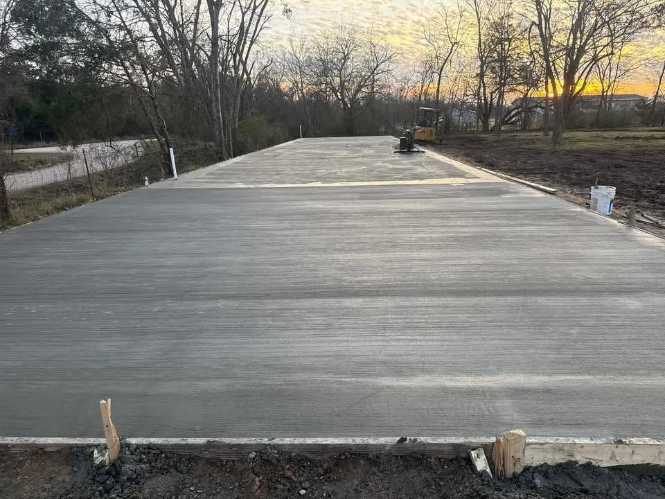 Freshly poured concrete slab at sunset, with forms and trees along a roadside lot.