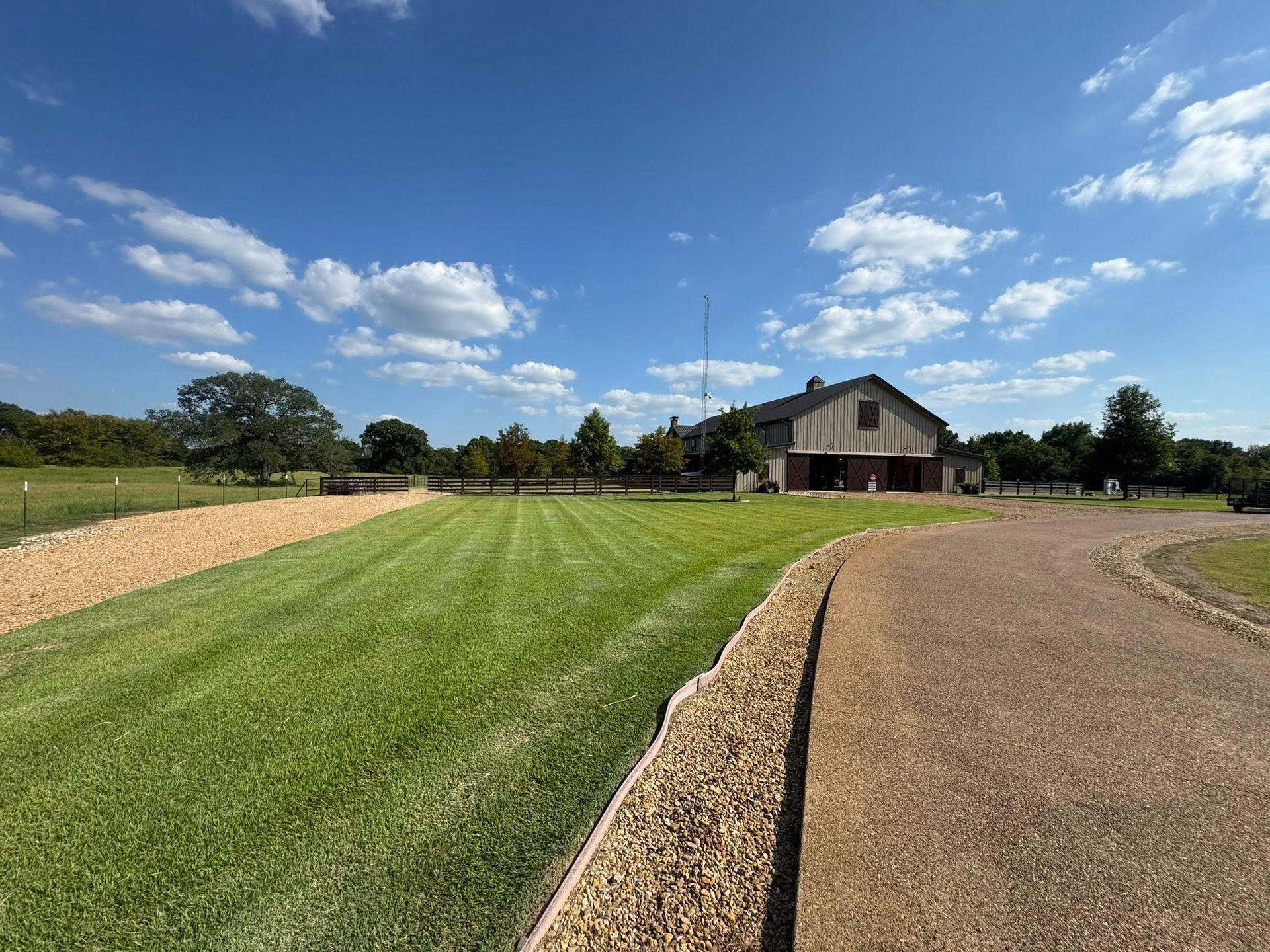 Wide gravel driveway leading to a brown barn across a green lawn under a blue sky