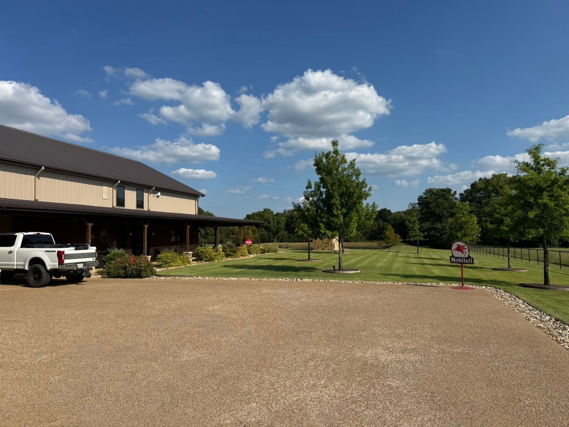 Gravel driveway beside a house, with a white pickup, trees, and a sunny blue sky with clouds.