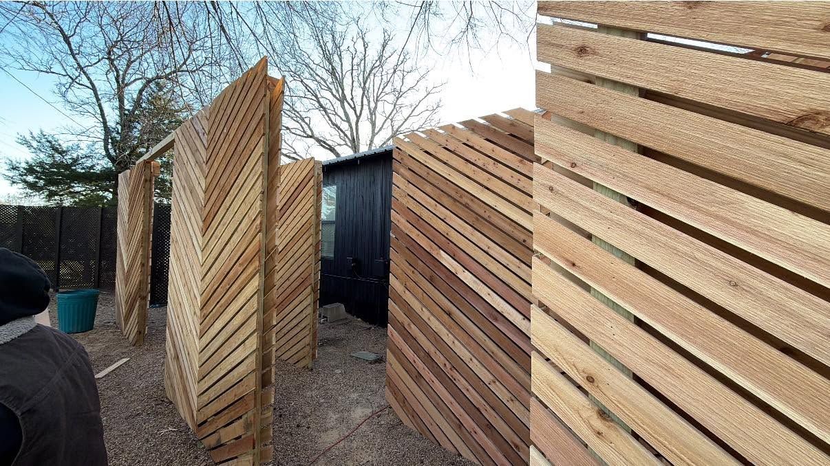 Wooden fence panels leaning in a yard, with a black gate and bare trees in the background.