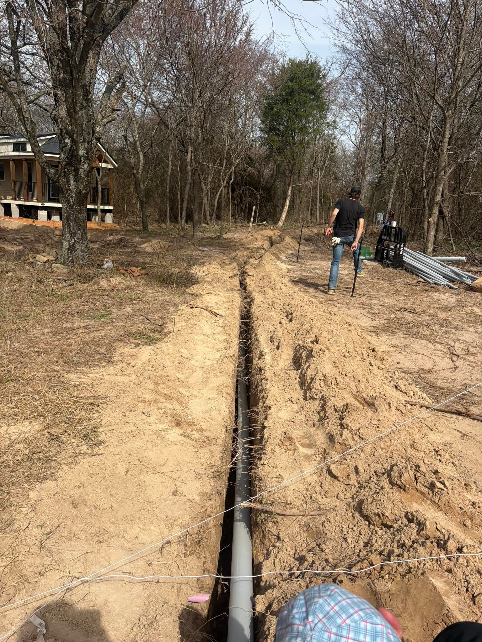 Trenched dirt path with a black pipe laid in the ground and two workers near a wooded area
