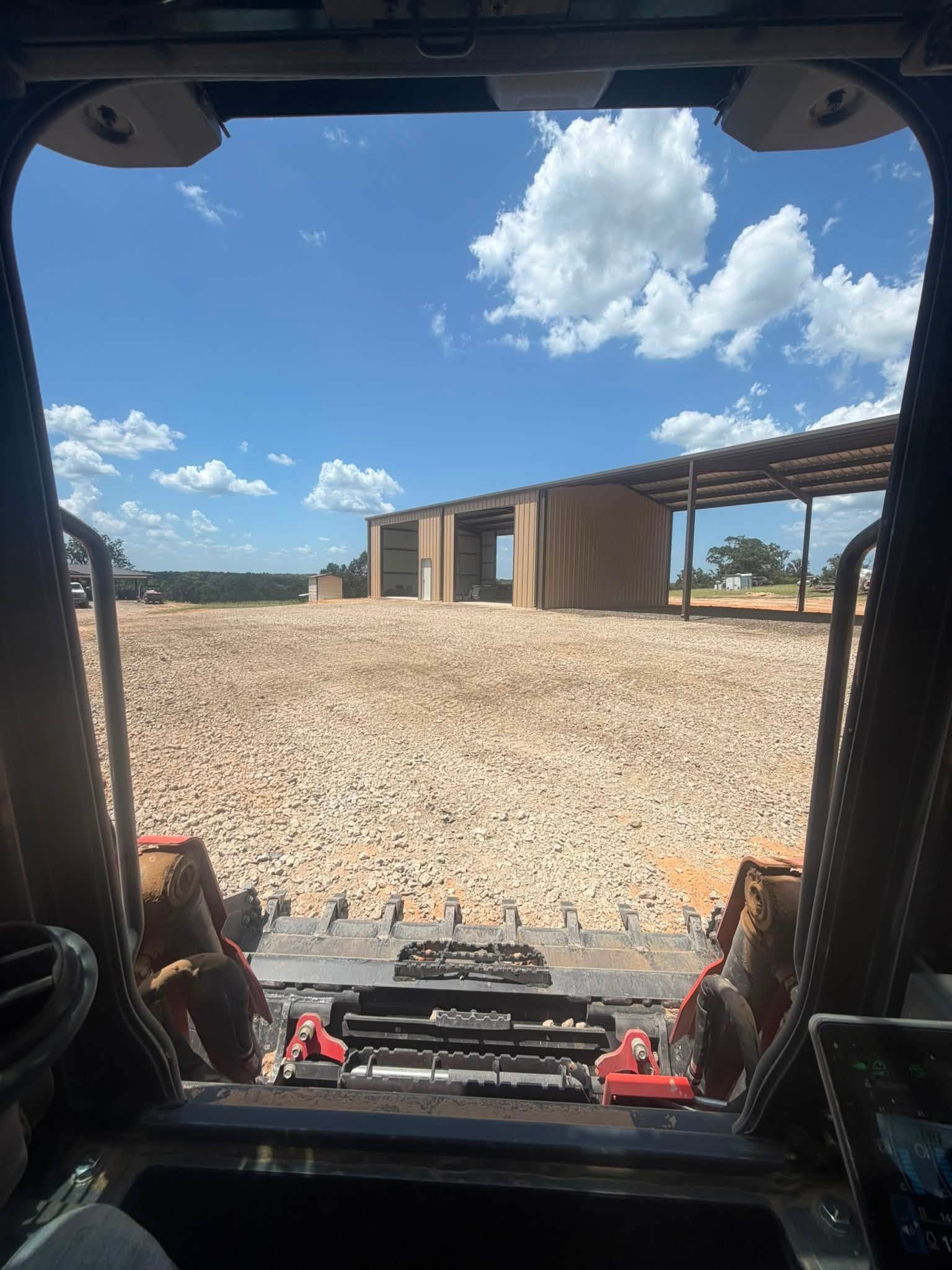 View from inside a construction vehicle across gravel to a modern building under a bright blue sky