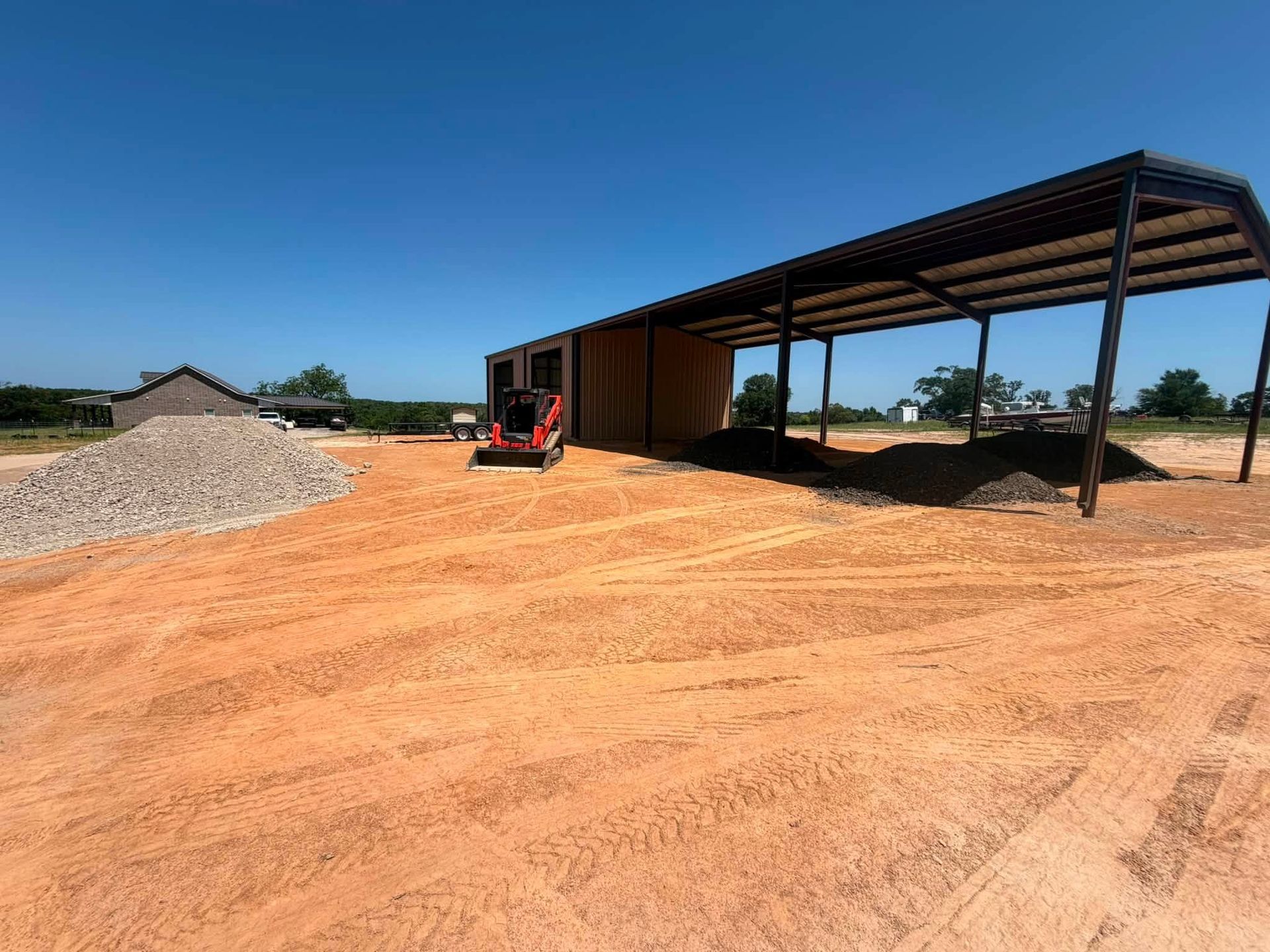 Large dirt lot with a gravel pile, open-sided shed, and a small excavator under a clear blue sky