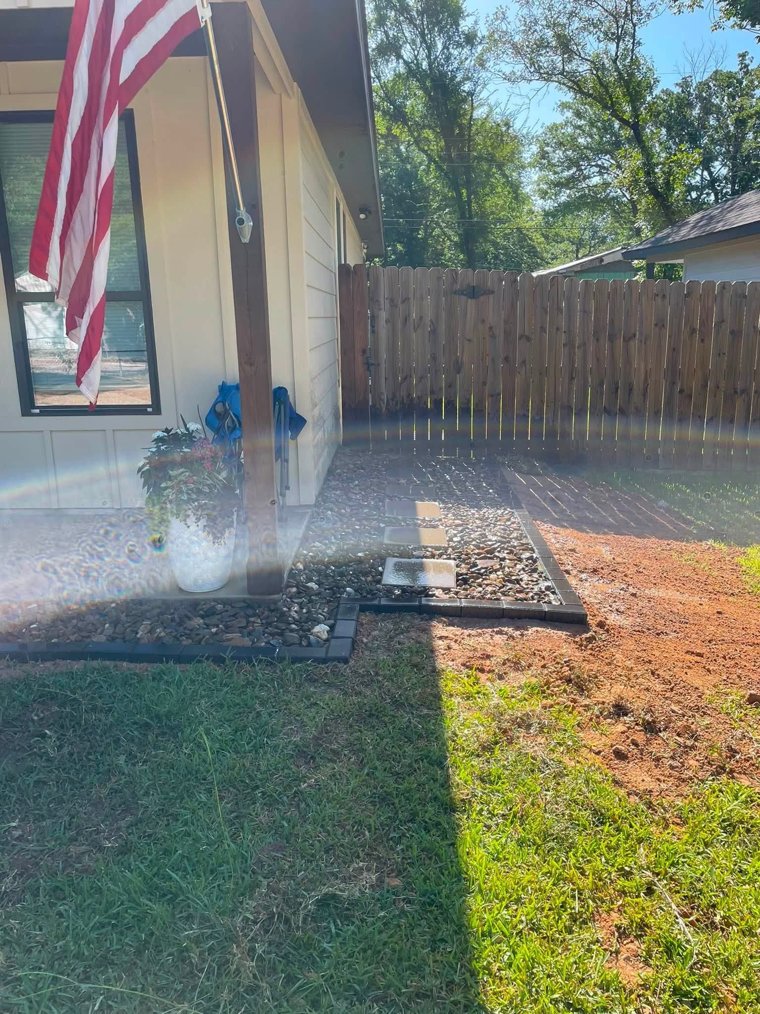 Patio sprinkler spraying water beside a house, with an American flag and fenced backyard in sunlight