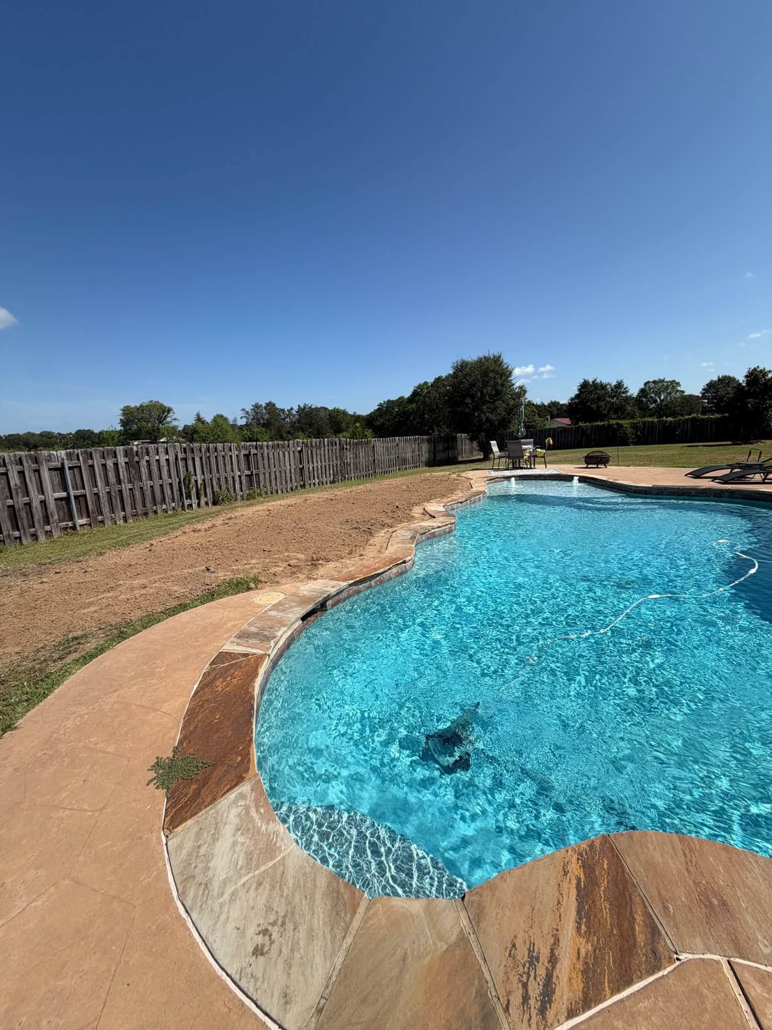 Curved backyard swimming pool with blue water, stone coping, and a fence under a clear sky