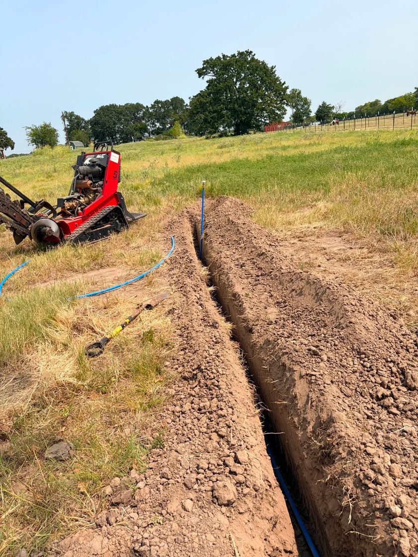 Red excavator digging a narrow trench through a grassy field under a clear sky.