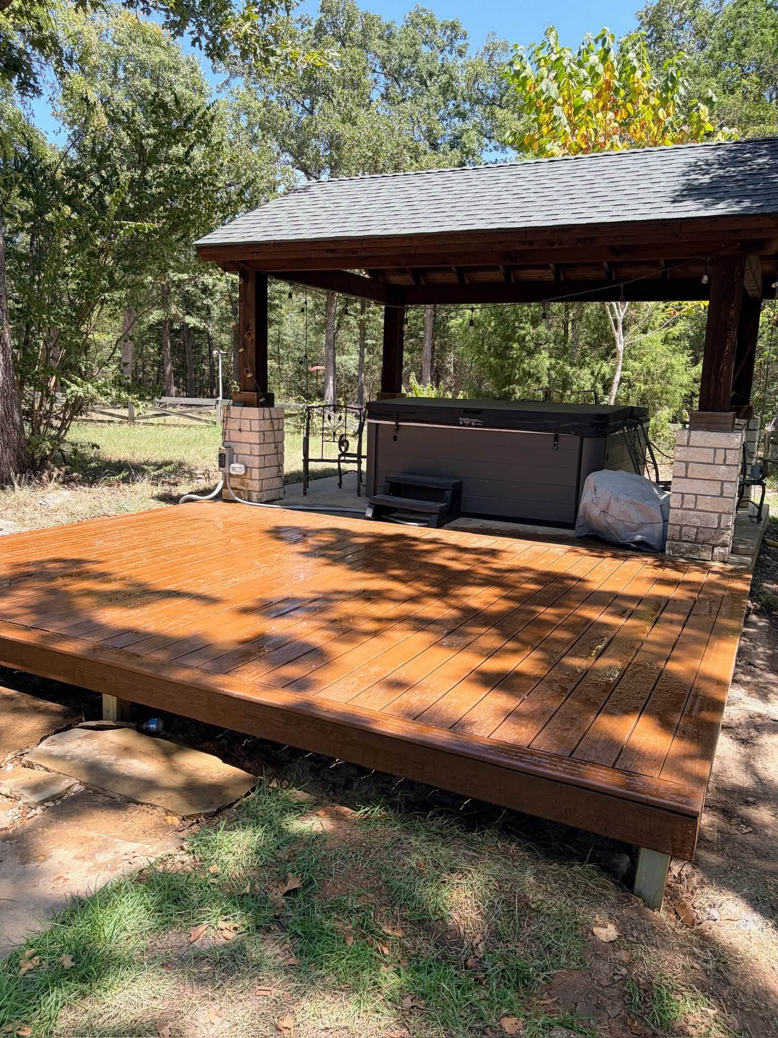 Wooden deck pavilion with a metal roof in a grassy backyard, surrounded by trees and shade.