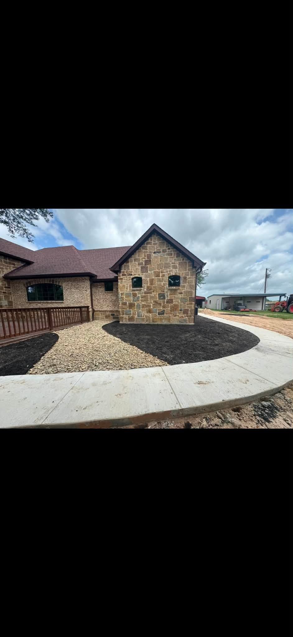 Stone house with a brown roof and a paved driveway under a cloudy sky.