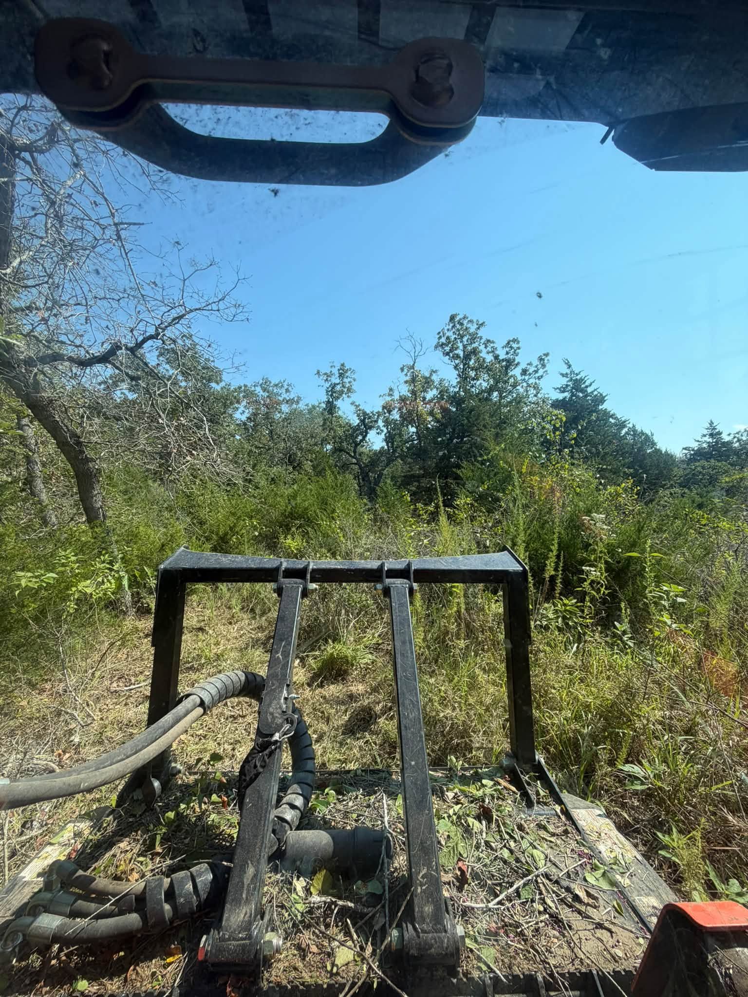 View from a vehicle over a metal front guard toward a muddy dirt track and dense green brush under a blue sky