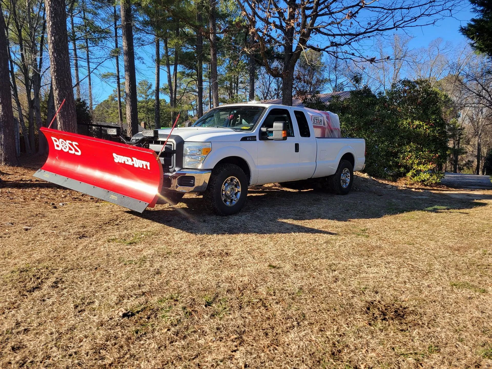 A white truck with a red snow plow on the back is parked in a dirt field.