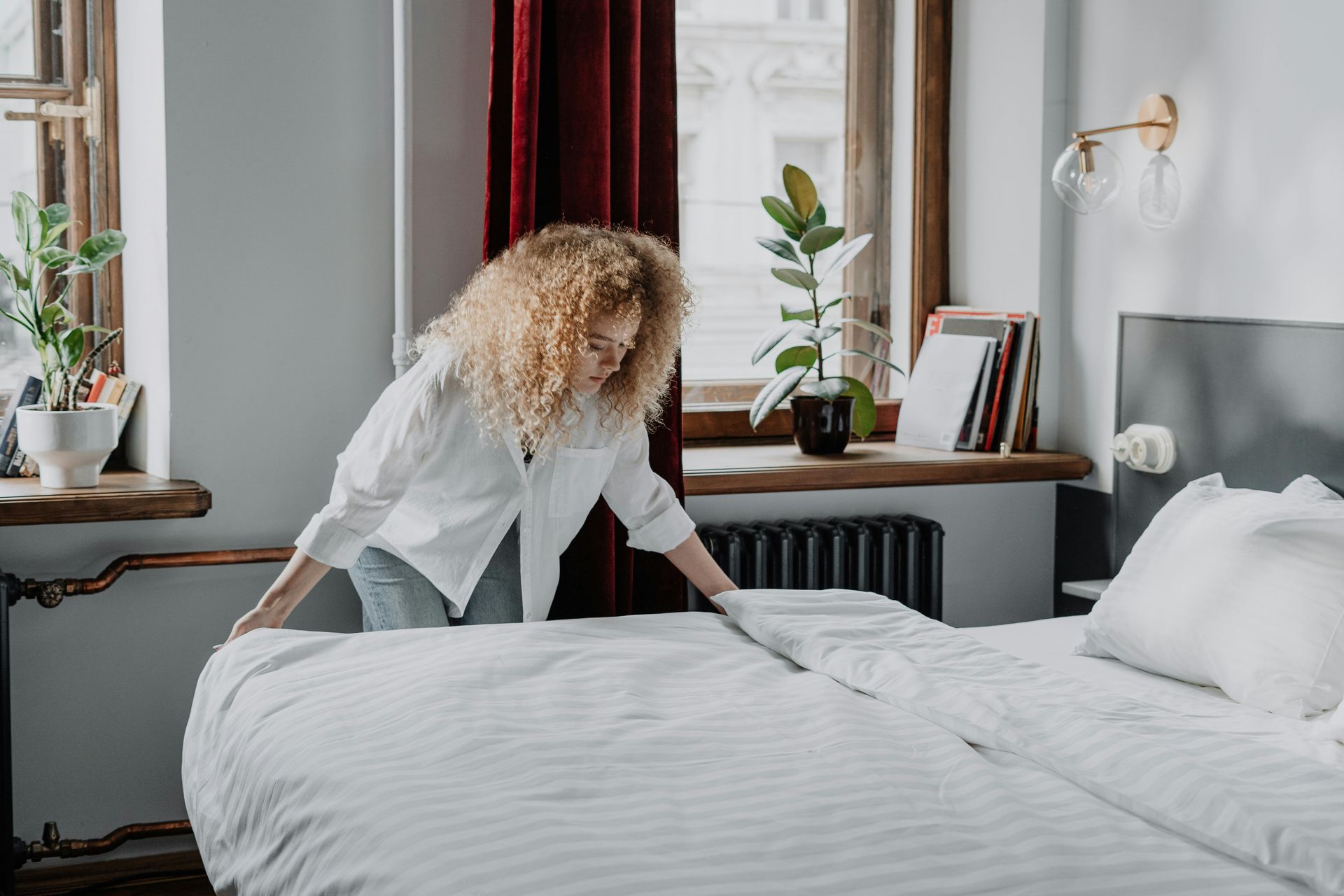 Woman making a bed in a bright bedroom, adjusting white bedding. Windows, plants, and a radiator are visible.