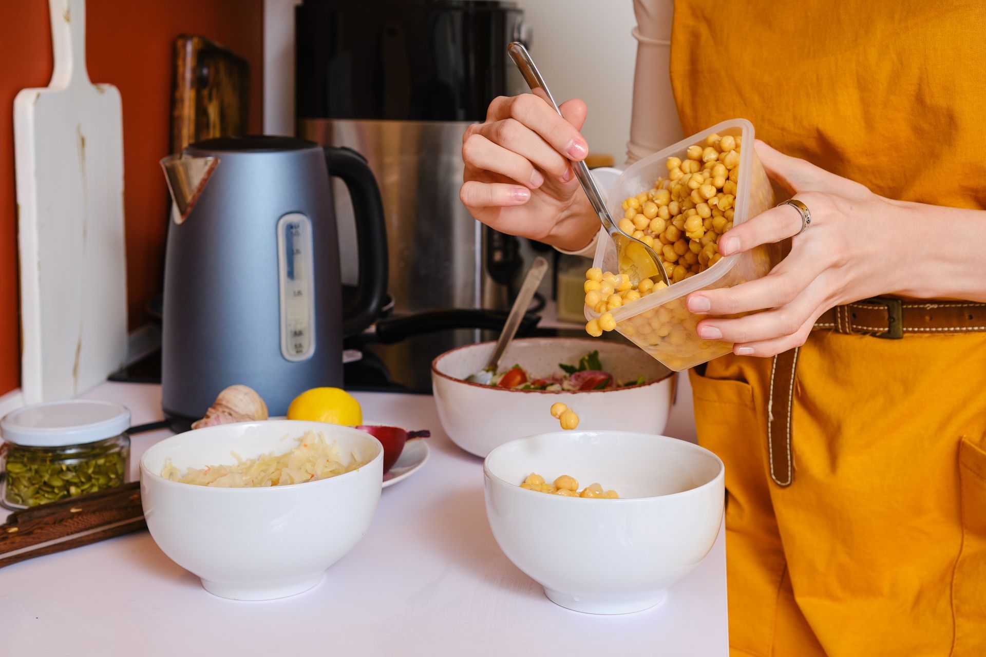 Woman in yellow apron preparing food, pouring chickpeas into a bowl in a kitchen setting.