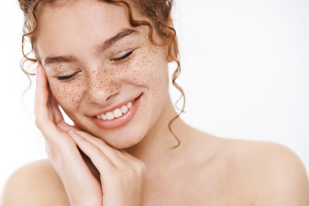 Smiling person with freckles touching their face against a white background