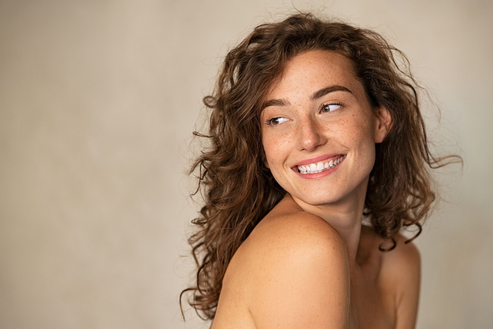 Woman with curly brown hair, smiling, looking to the side, against a neutral background.