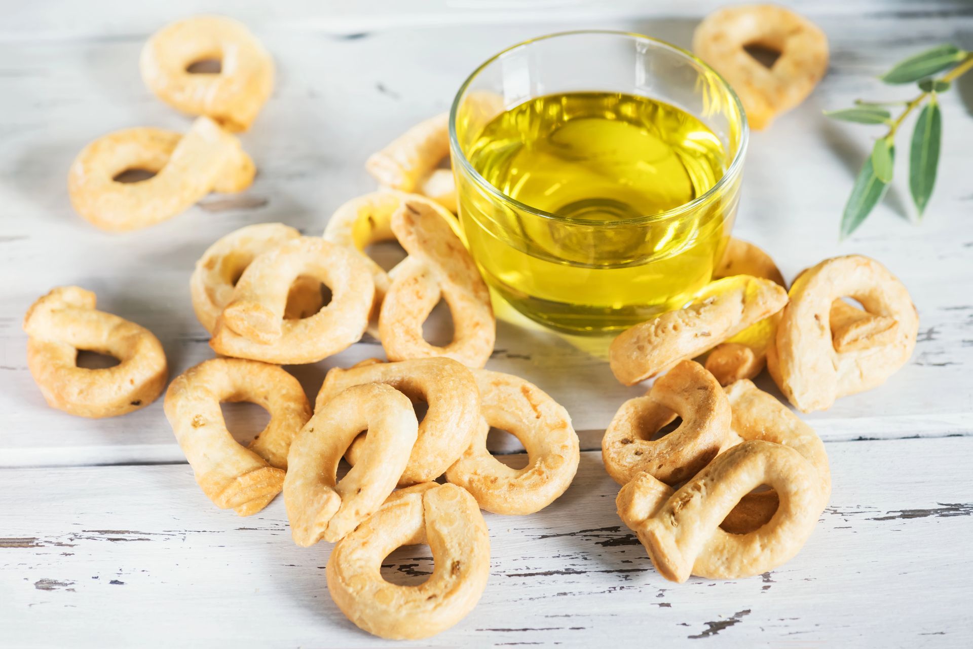 Un bicchiere di olio d'oliva circondato da piccoli cracker a forma di anello su una superficie di legno bianco.